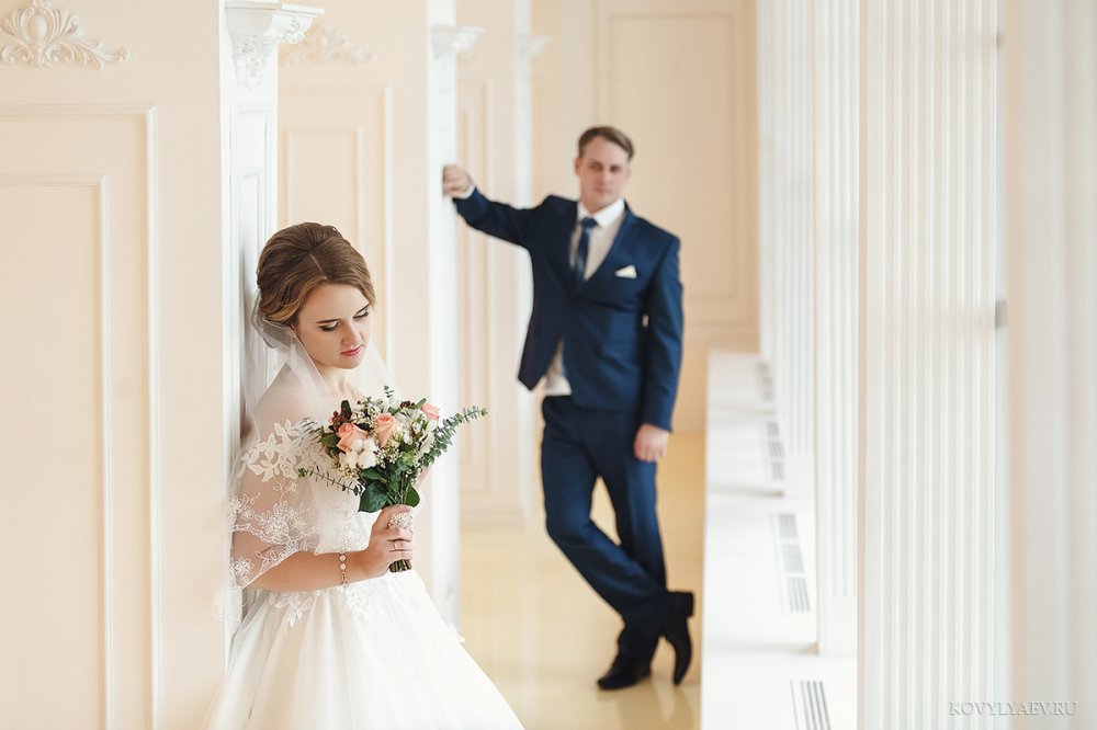 Невеста на переднем плане, жених на заднем стоит, облокотившись на колонну.